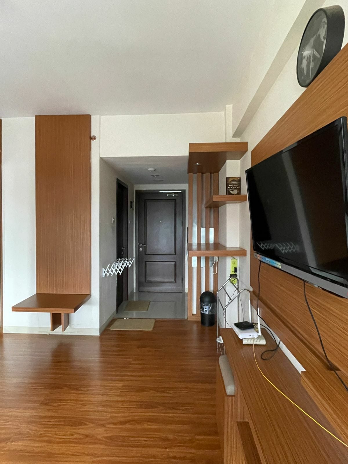 An entrance area features wooden accents and warm tones. A wall-mounted television is positioned above a low shelf, while a small table extends from the wall. The space is illuminated by natural light, leading to a door that opens into the private room.