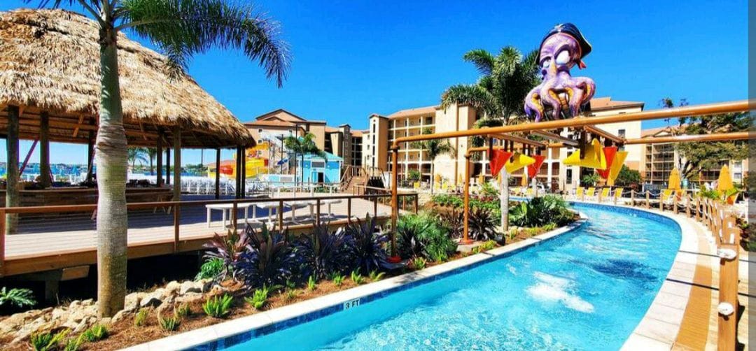 A vibrant water park is featured, showcasing a lazy river surrounded by lush tropical plants. A colorful octopus structure decorates the area, with a thatched-roof gazebo visible nearby. Bright sunshine reflects off the water, creating a lively atmosphere suitable for families.