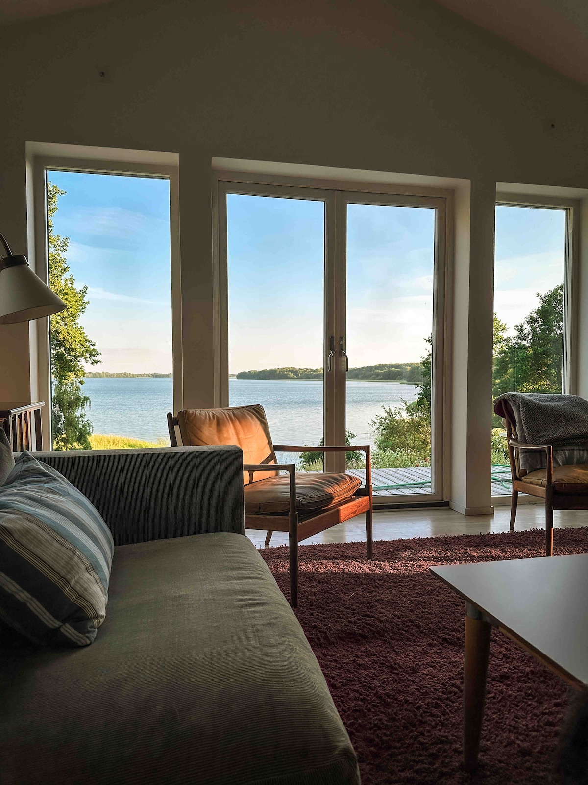 Large windows offer a panoramic view of the serene lake, allowing natural light to fill the living space. Comfortable seating is arranged for relaxation, with a soft rug underfoot complementing the inviting atmosphere.