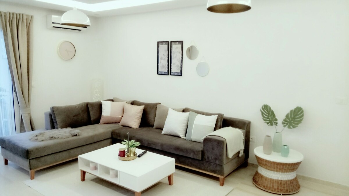 A modern living area features a plush sectional sofa adorned with decorative pillows in soft hues. A white coffee table sits at the center, accompanied by a round wicker side table. Wall art adds subtle character, while natural light enters through the window with light curtains.