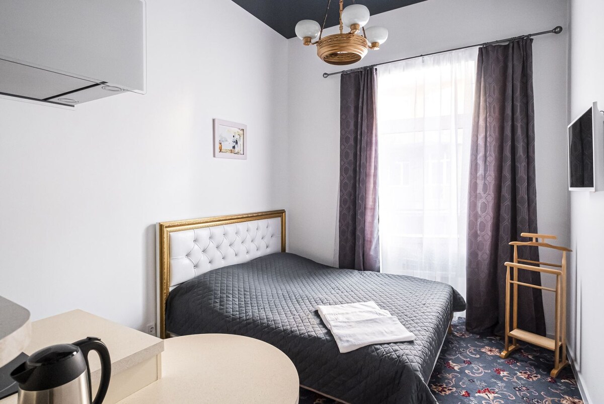 A spacious sleeping area is featured, with a large bed adorned in dark linens. Natural light enters through a tall window covered by sheer curtains, while a simple wooden clothes rack adds functionality. A small kitchenette area is visible in the background.