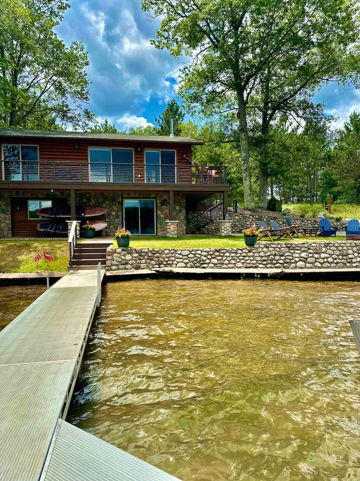 A lakeside cottage is visible, featuring a stone and wood exterior. A dock extends towards the water, while a spacious outdoor area includes a patio and seating options. Lush greenery and flower planters add a natural touch, set against a backdrop of trees and a partly cloudy sky.
