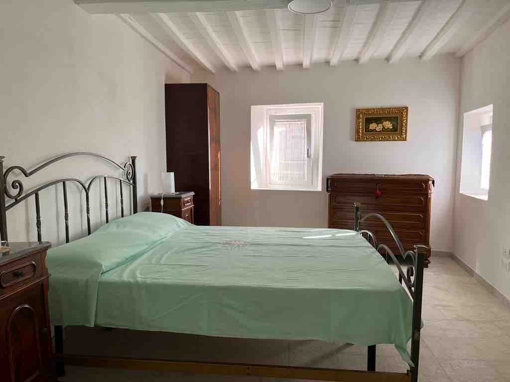 A spacious bedroom is furnished with a metal bed and light green bedding. Wooden furniture, including a wardrobe and a chest, complements the light-colored walls. Natural light enters through two small windows, highlighting the room's simple design and architecture.