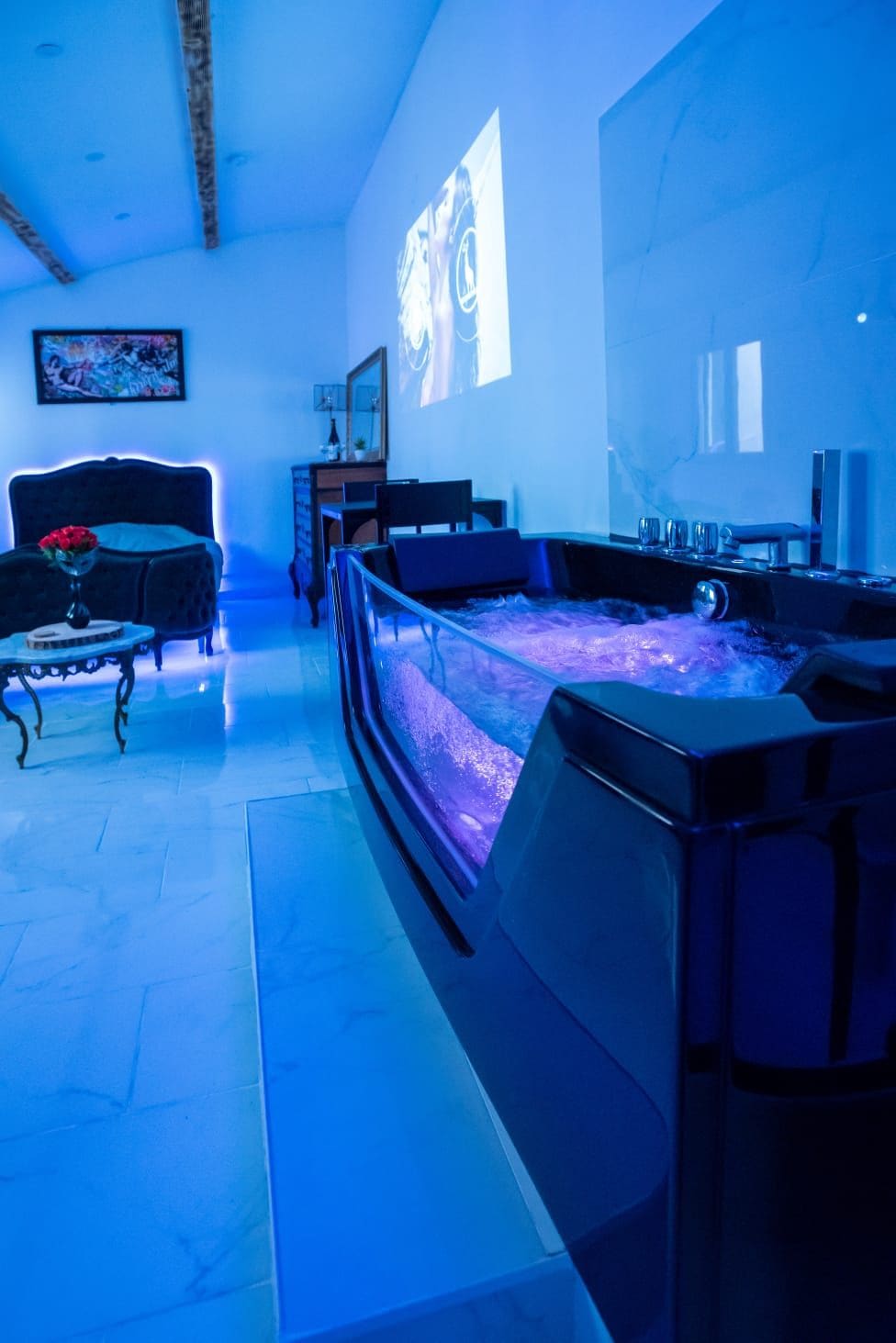 A stylish and tranquil space features a modern balneotherapy tub with soft blue lighting. A comfortable seating area is visible, adorned with a small table holding a vase of red flowers. The ambient atmosphere is complemented by the subtle glow from decorative elements.