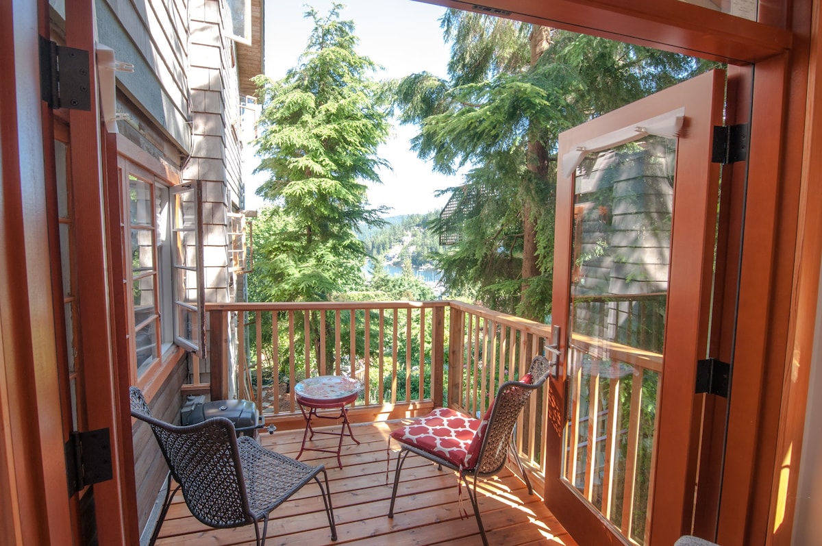A private deck is depicted with two chairs, one featuring a patterned cushion. A small round table is positioned between them. Lush greenery surrounds the space, providing views of the nearby water, contributing to a serene outdoor setting.