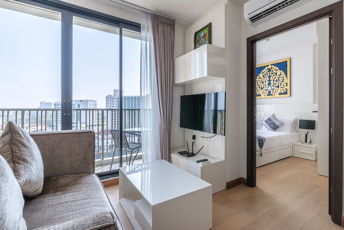 A stylish living area features a comfortable sofa and a white coffee table, set against large glass windows that provide ample natural light. A wall-mounted television adds modern convenience, while a glimpse of a cozy bedroom can be seen through the open door.