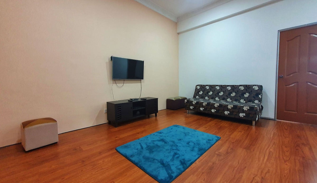 A living area is presented with a flat-screen television mounted on the wall. A dark sofa bed adorned with floral patterns faces a low black media console. A soft blue rug adds color to the warm, wooden flooring, while a closed door stands on one side.