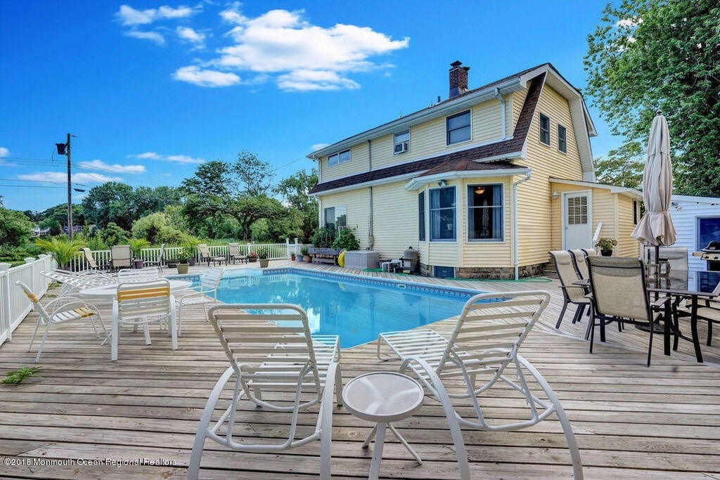 A spacious pool deck surrounds a clear blue pool, featuring multiple lounge chairs and dining tables. The house is painted yellow with large windows, surrounded by greenery. The area is brightened by a blue sky dotted with white clouds, creating a welcoming outdoor setting.