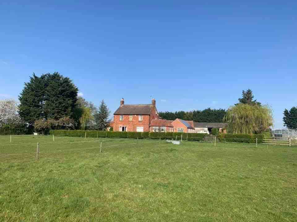 A charming farmhouse is set among green fields, surrounded by a low hedge. The two-story red brick structure is complemented by a single-story brick extension, with trees visible in the background under a clear blue sky.