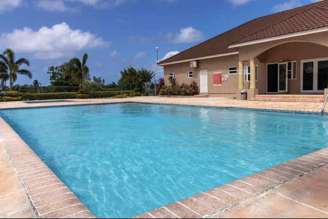 A sparkling blue pool is surrounded by well-maintained landscaping and a clear sky. A building with large windows and a sloped roof is visible in the background, offering easy access to the pool area.