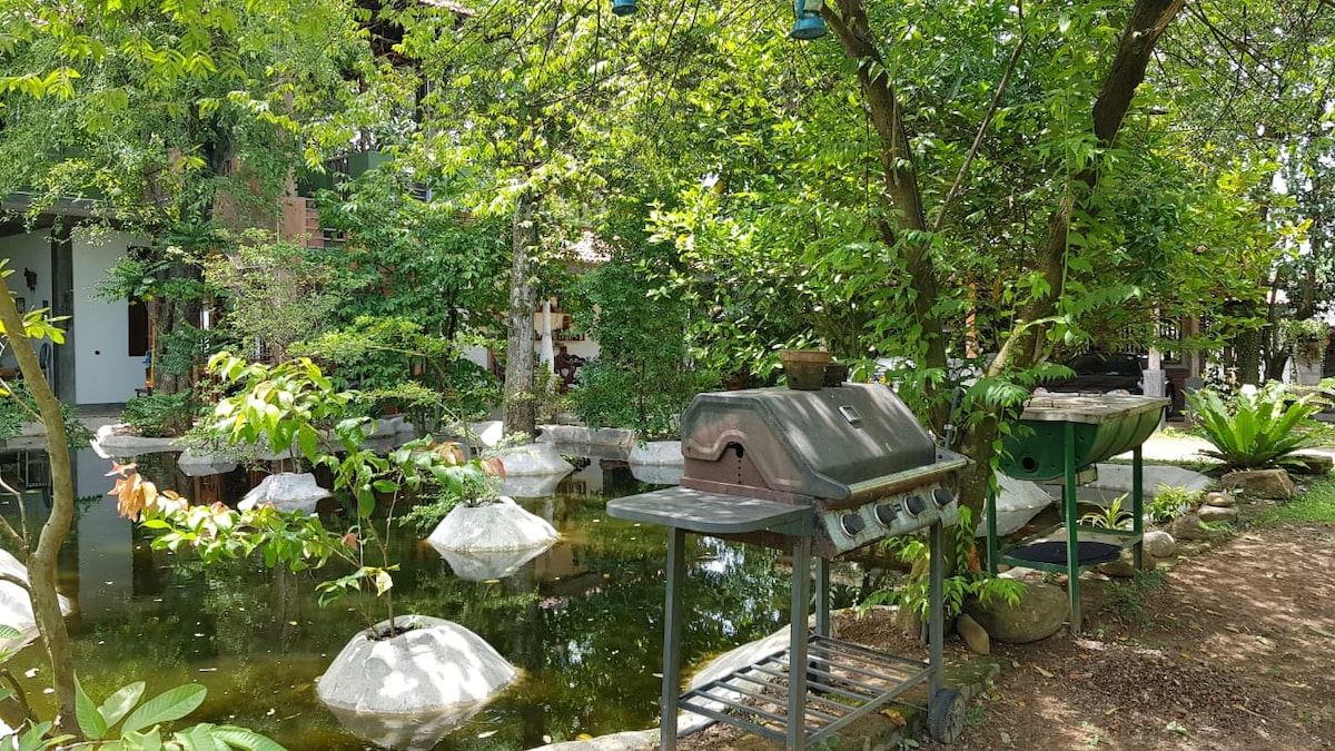 A serene outdoor space is depicted with a pond surrounded by lush greenery and large rocks. A barbecue grill and prep area sit nearby, inviting the opportunity for outdoor cooking. The tranquil setting features various trees and plants, enhancing the natural ambiance.