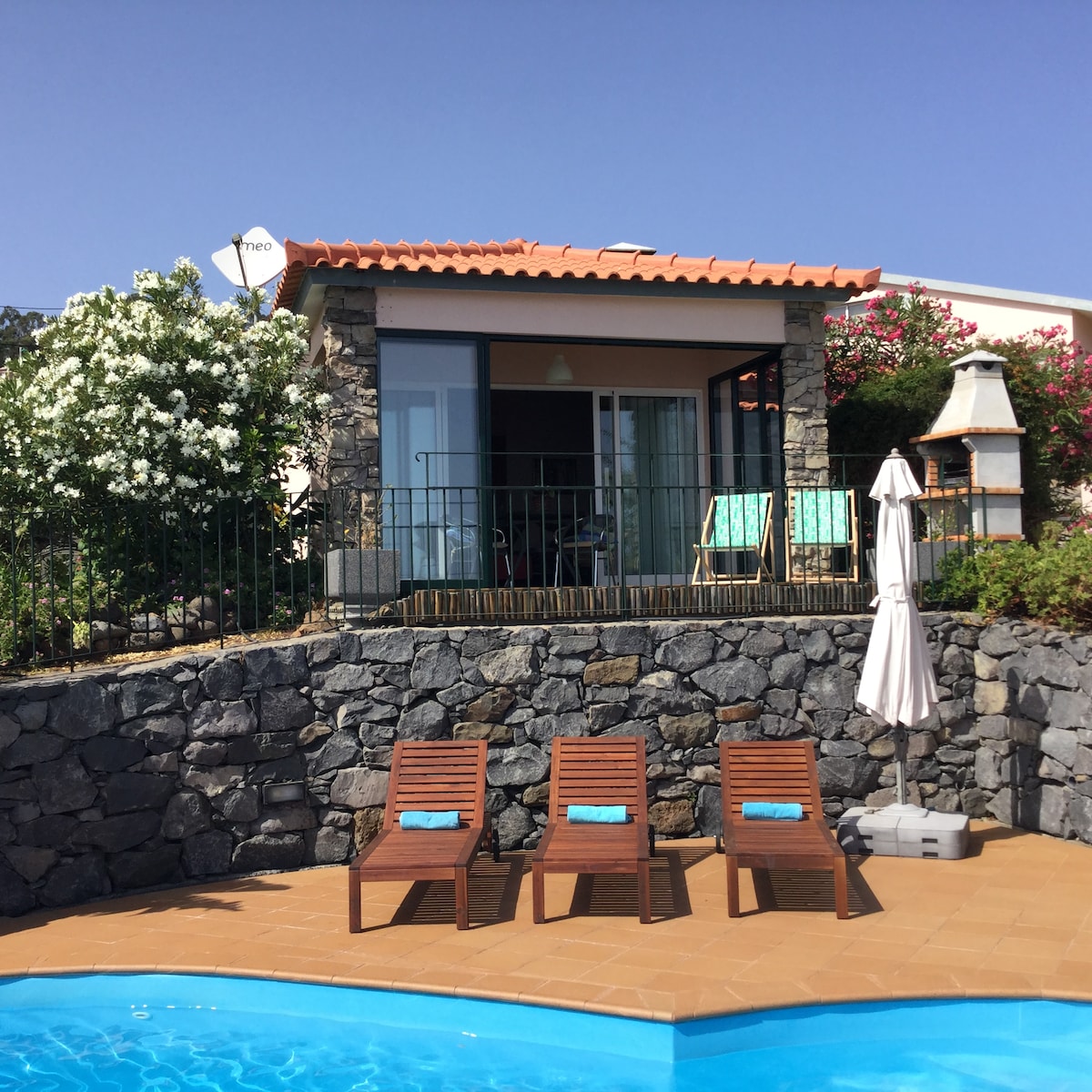 A cozy cottage is set amongst vibrant green foliage, with a stone wall enclosing a sparkling swimming pool. Four wooden loungers with blue cushions are arranged around the pool, while a sun umbrella offers shade. The glass-fronted cottage features a welcoming patio area.