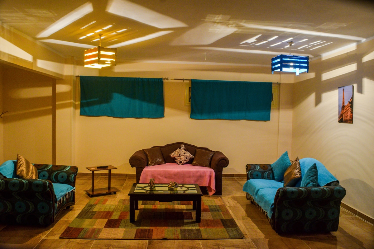 A spacious living area is furnished with a brown sofa and two vibrant blue chairs, complemented by colorful overhead lighting. Decorative throw pillows are placed on the seating, while a coffee table is positioned in the center, atop a colorful area rug.