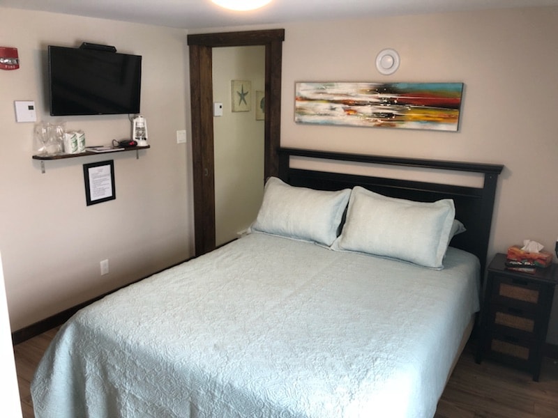 A comfortable sleeping area features a queen size bed dressed in light blue bedding. An abstract piece of wall art adds a touch of color above the headboard. A flat-screen TV is mounted on the wall opposite the bed, while a nightstand holds various essentials.