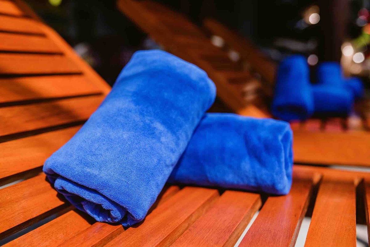 Two rolled blue towels are arranged neatly on a wooden lounger. The sunlight reflects off the smooth wood, creating a warm, inviting atmosphere for relaxation.