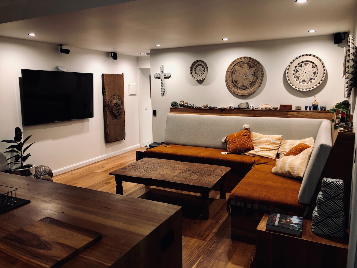 A cozy living area features a large sectional sofa with warm-colored cushions, positioned around a rustic wooden coffee table. Wall decor includes woven art pieces and a cross, while a flat-screen TV is mounted nearby. Warm lighting contributes to a relaxing environment.