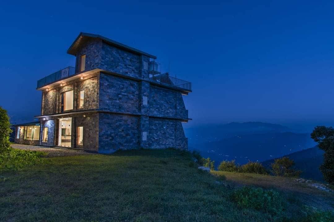 A modern multi-story cottage is showcased against a twilight backdrop, featuring a stone exterior and large windows that illuminate the interior. The surrounding landscape offers a serene view of distant hills and a softly lit sky.