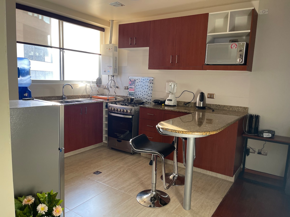 An equipped kitchen is featured, showcasing light wood cabinetry and a granite countertop. A stainless steel stove and oven are positioned beside a built-in microwave. A glass-top table with a black stool is included, with a large window providing ample natural light.