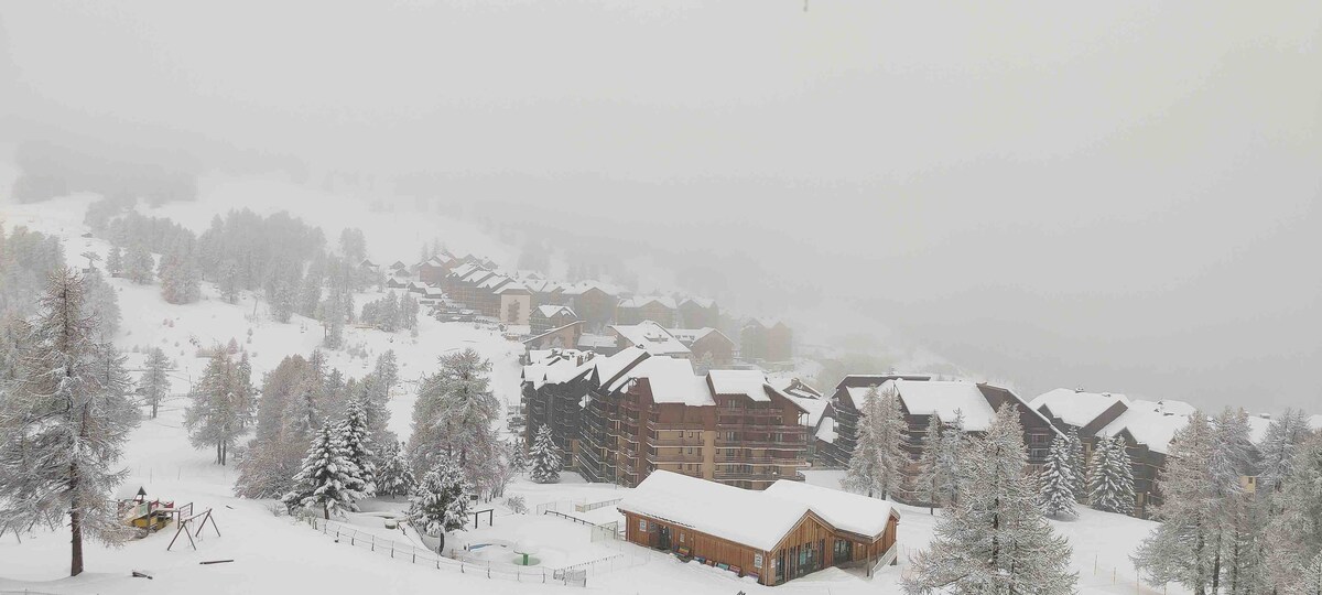 A panoramic winter view shows snow-covered buildings nestled among trees. The landscape is blanketed in white, with a soft mist enveloping the surroundings. A children's swing set is visible in the foreground, emphasizing a serene and tranquil atmosphere.