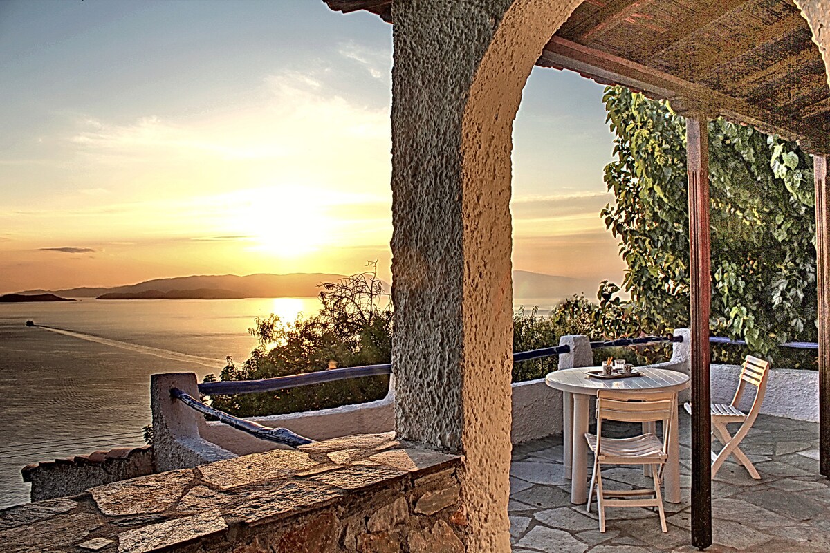 A shaded outdoor terrace features a round table and white chairs, offering unobstructed views of the sunset over the sea. The stonework and foliage create a serene area for relaxation, complementing the natural beauty of the surrounding landscape.