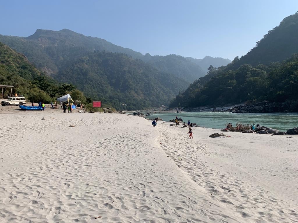 A sandy beach area is bordered by a river, with gentle waves lapping at the shore. Lush green mountains rise in the background, and groups of people can be seen enjoying the outdoors. A few tents and vehicles are placed along the beach.