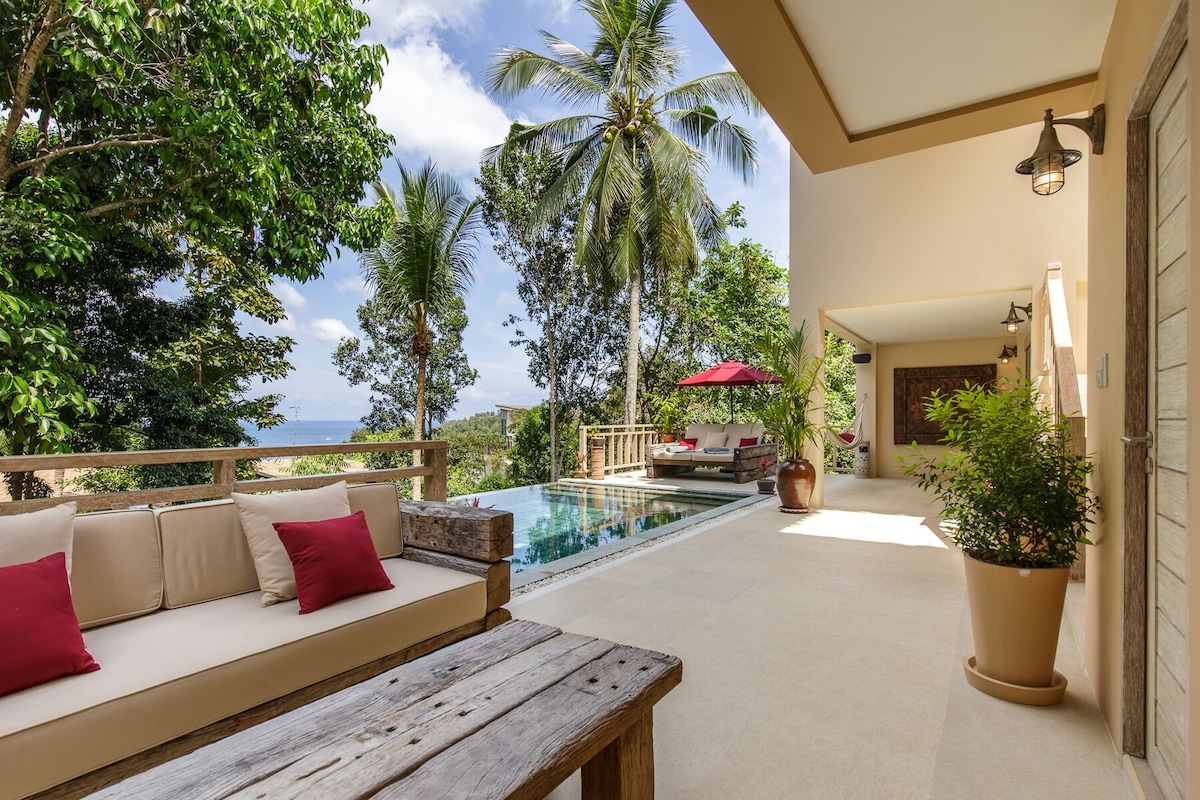 An outdoor lounge area features a spacious beige couch adorned with red cushions, positioned near a tranquil infinity pool. Lush greenery surrounds the space, with palm trees swaying gently in the breeze. A sun-lounge area with a red parasol is visible in the background.