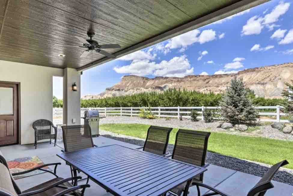 A covered patio area is furnished with a dining table and several chairs, providing comfortable seating. A view of the landscaped yard is framed by a white fence, with mountains visible in the distance under a partly cloudy sky.