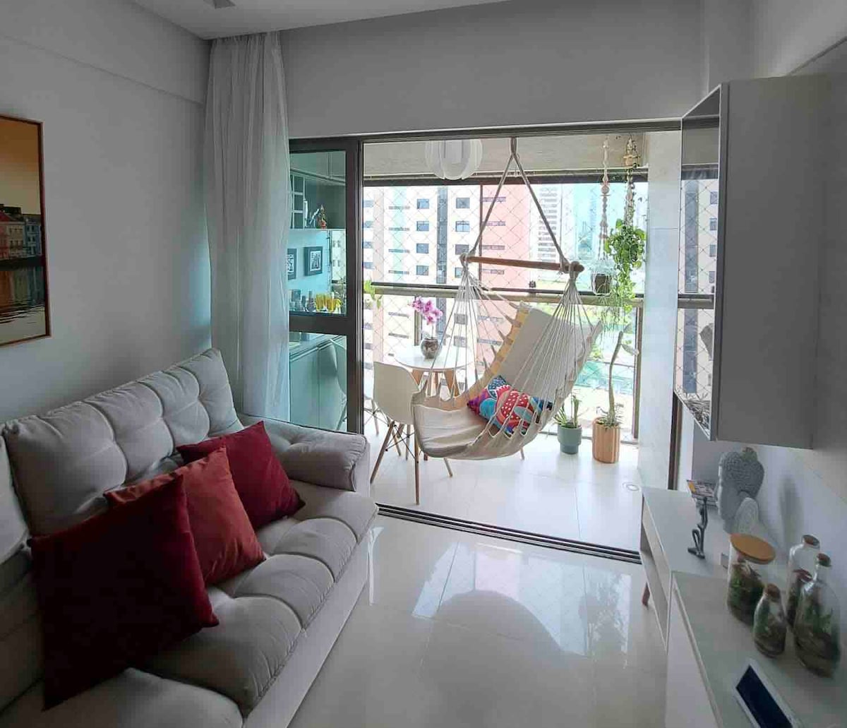 A cozy living space features a light-colored sectional sofa adorned with red and pink cushions. A uniquely designed hammock chair is positioned near large glass doors, leading to a terrace lined with greenery. Natural light floods the room, creating an inviting atmosphere.