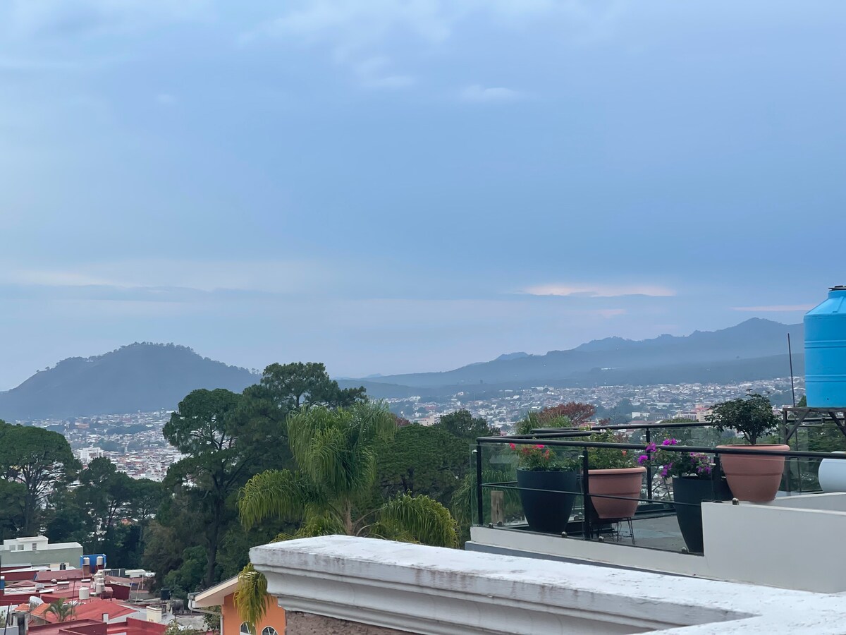A tranquil terrace offers scenic views of rolling hills and scattered urban landscapes. Potted plants line the space, adding a touch of greenery against the backdrop of the horizon, with soft clouds visible above the distant mountains.
