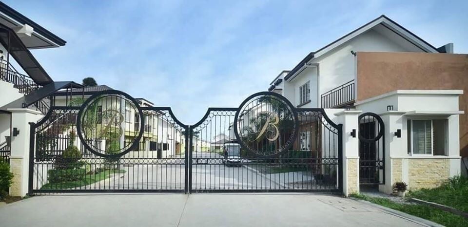 A gated entrance is showcased, featuring ornate iron gates with a circular design. The pathway leads into a private community flanked by well-maintained homes, highlighted by a clear sky above and landscaped elements at the entrance.