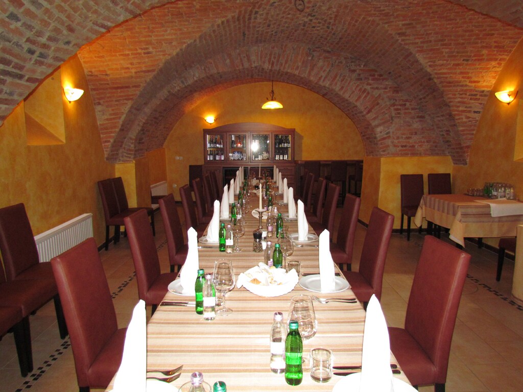 A spacious dining area features a long table set with white linens, neatly arranged tableware, and green bottled drinks. Rows of brown upholstered chairs line the table, complemented by arched brick ceilings and warm lighting that enhances the room's inviting atmosphere.