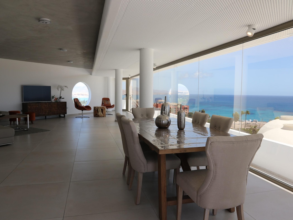 A dining area features a large wooden table surrounded by upholstered chairs. Floor-to-ceiling windows provide expansive views of the sea and let in ample natural light. A cozy seating area is visible in the background, arranged for relaxation and entertainment.