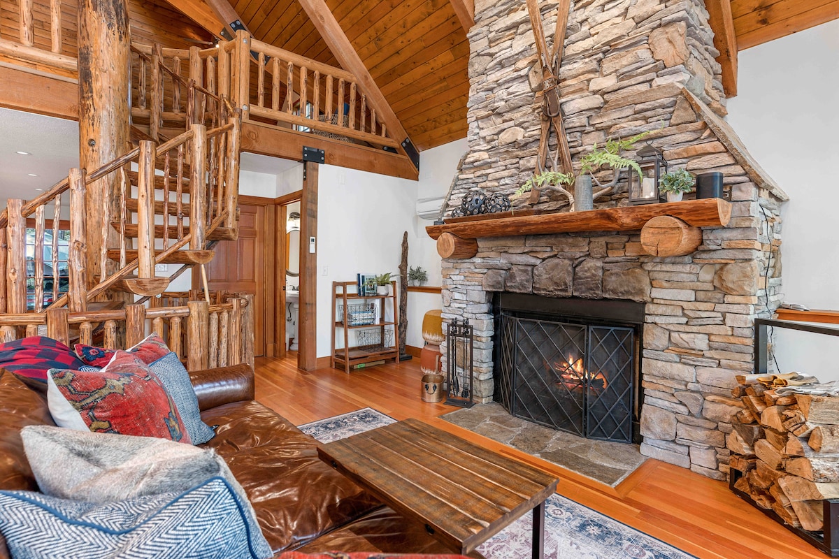 A spacious living area features a prominent stone fireplace with a wood-burning fire. Rustic wood accents are visible throughout, including a spiral staircase leading to the upper level. Comfortable seating is arranged around a wooden coffee table, offering a cozy gathering space.
