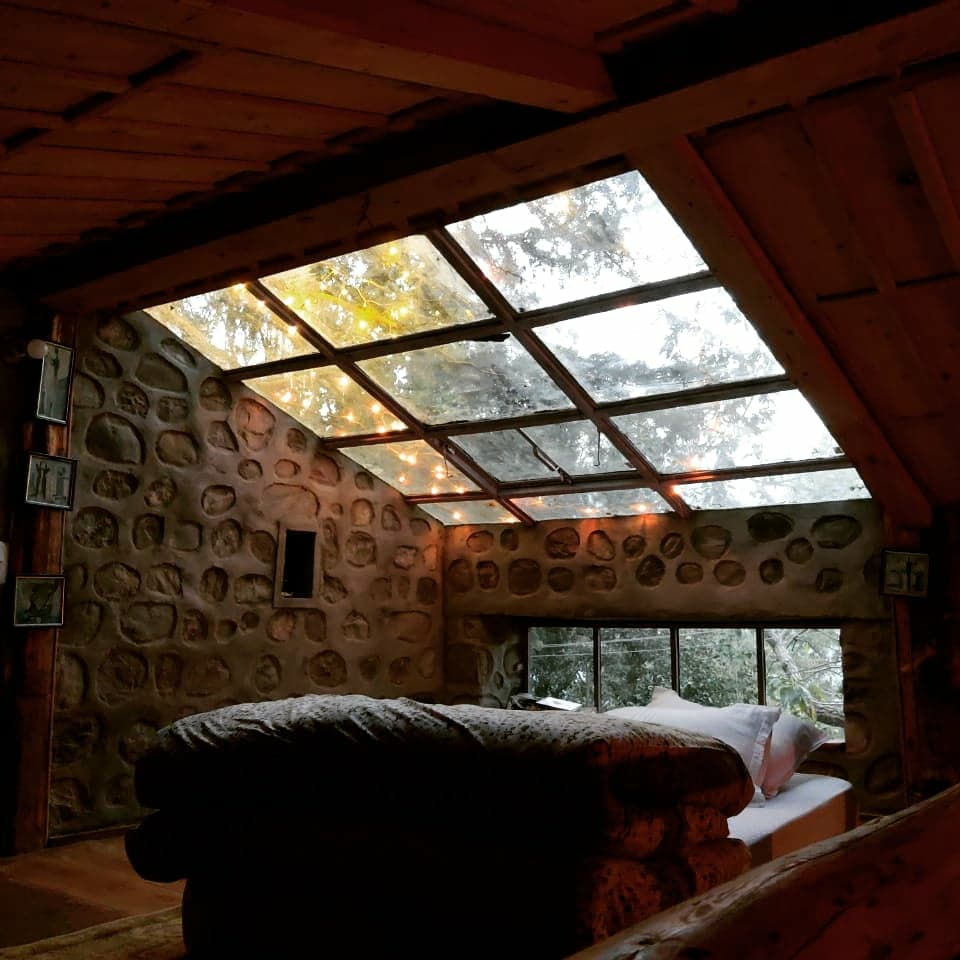 A cozy interior features a spacious bed covered with a warm blanket, surrounded by natural stone walls. Large windows, including a skylight, allow for ample natural light and views of the surrounding trees, enhancing the serene atmosphere of the hut.