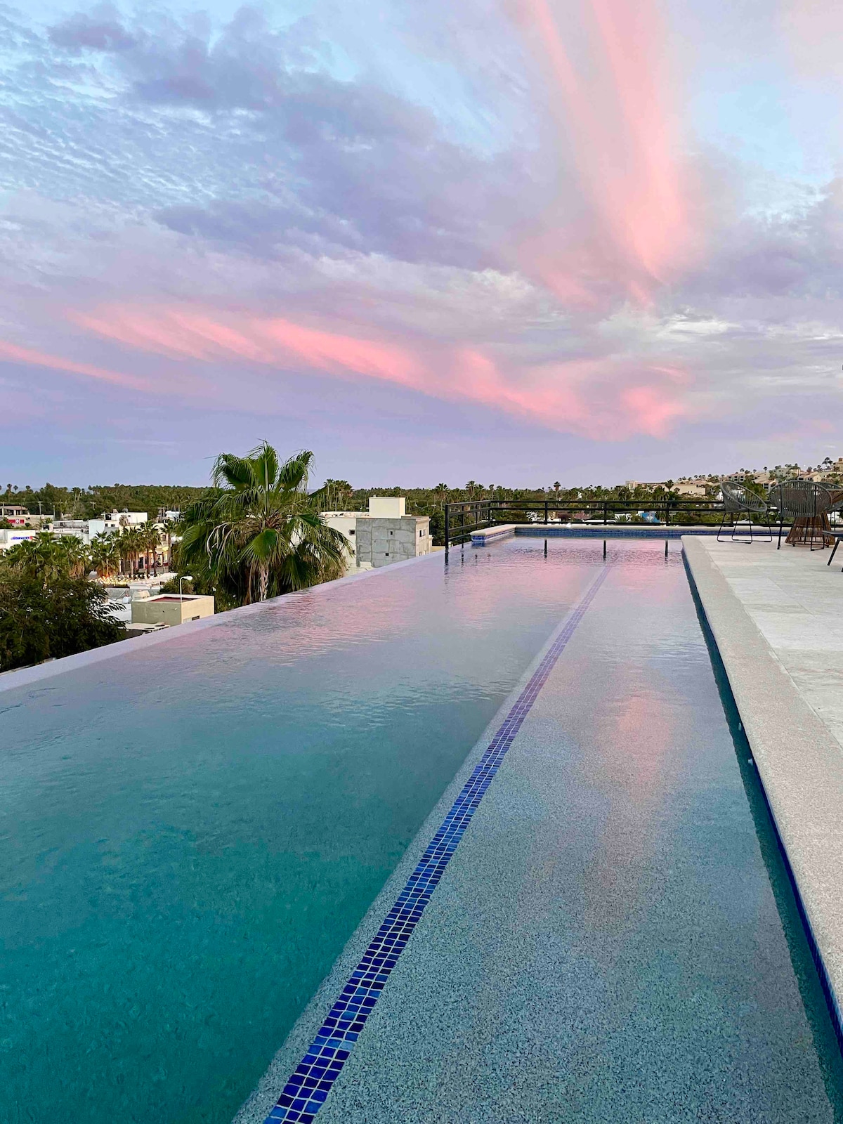 An infinity pool extends towards the horizon, gently reflecting the pastel hues of a sunset sky. The pool's edges blend seamlessly into the landscape, surrounded by palm trees and tranquil views of nearby hills.