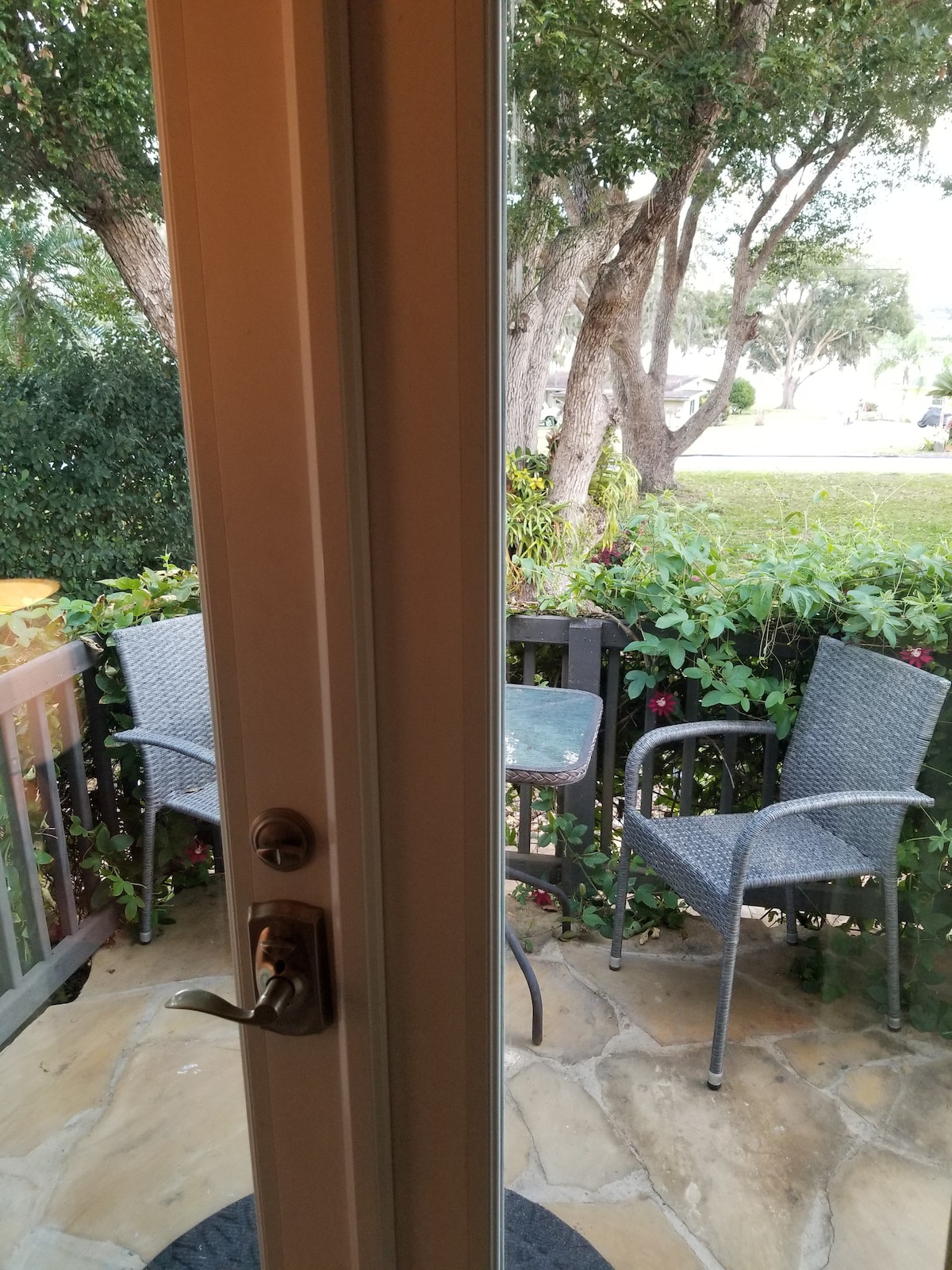 A small veranda is visible through a glass door, featuring two wicker chairs and a table. Lush greenery surrounds the area, providing a natural setting. The stone floor enhances the outdoor ambiance, and sunlight illuminates the space, creating a welcoming environment.
