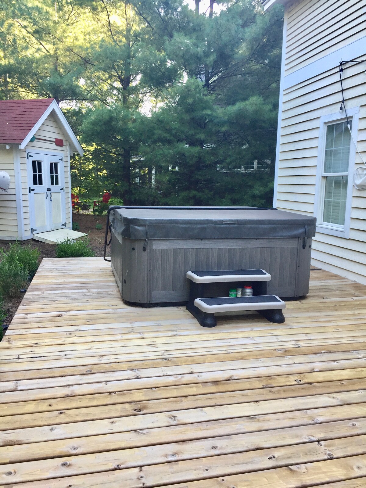 A hot tub is positioned on a wooden deck, surrounded by greenery. A set of black steps leads up to the tub, which is covered and situated near a playhouse. Soft sunlight filters through the trees, creating a peaceful outdoor setting.