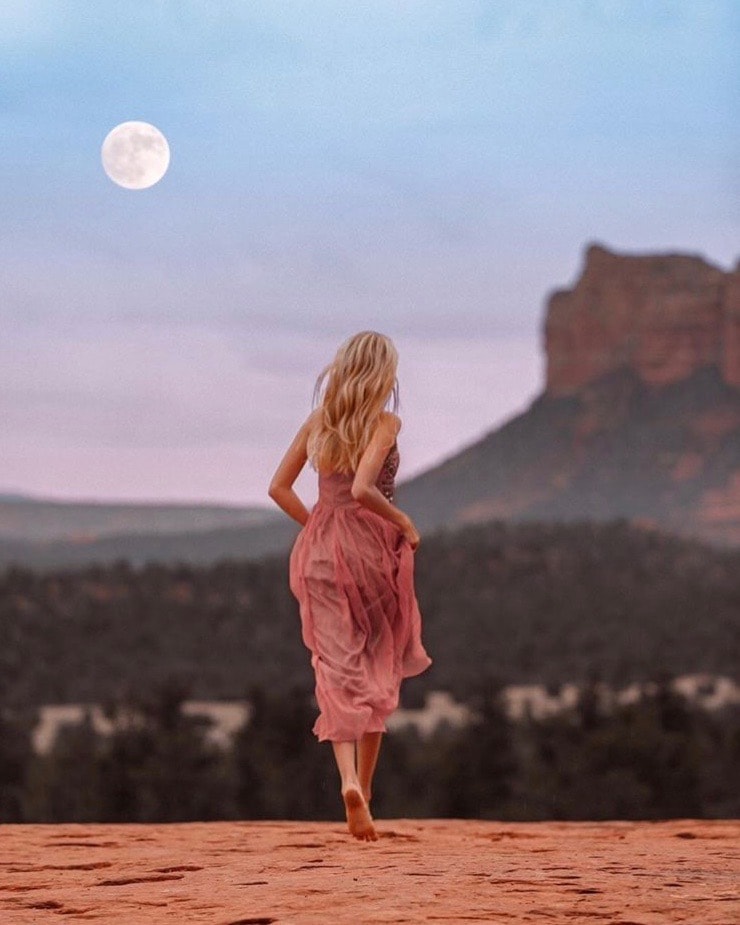 A person is seen walking barefoot across a sandy landscape under a large moon. Rolling hills and a prominent rock formation are present in the background, contrasted against a soft, pastel sky.