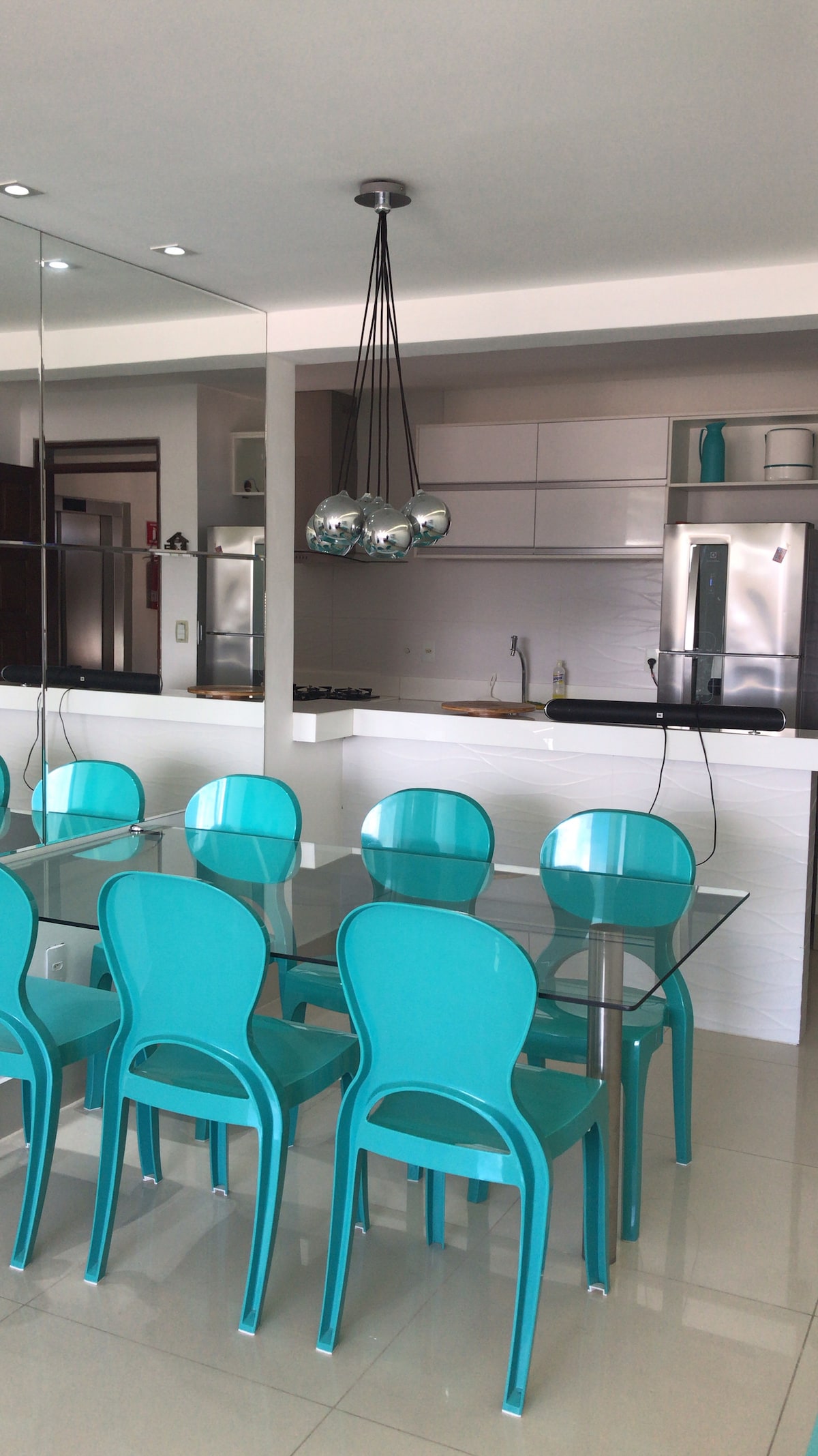 A modern dining area is presented with a glass table surrounded by six turquoise chairs. Above the table, a unique pendant light with three spherical bulbs provides illumination. The sleek kitchen layout is visible in the background, featuring stainless steel appliances and minimalist cabinetry.