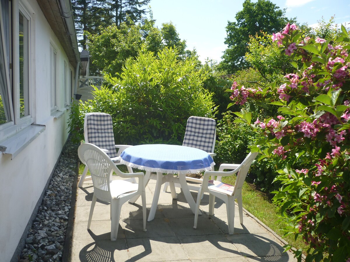 A private outdoor terrace features a circular table with a blue tablecloth surrounded by four chairs. Lush greenery is present, including flowering plants and shrubs, creating a serene setting. Sunlight casts a warm glow over the space, enhancing its natural appeal.