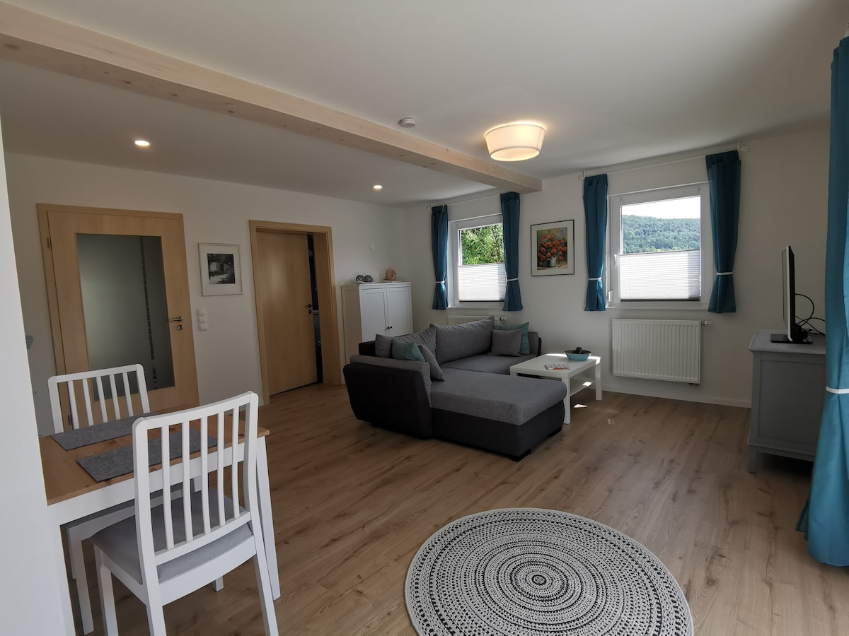 The living area features a spacious seating arrangement with a gray sofa and a neutral-toned rug. A dining table with two chairs is positioned nearby, while two windows provide natural light and a view of the outside greenery. Light-colored walls complement the modern decor.