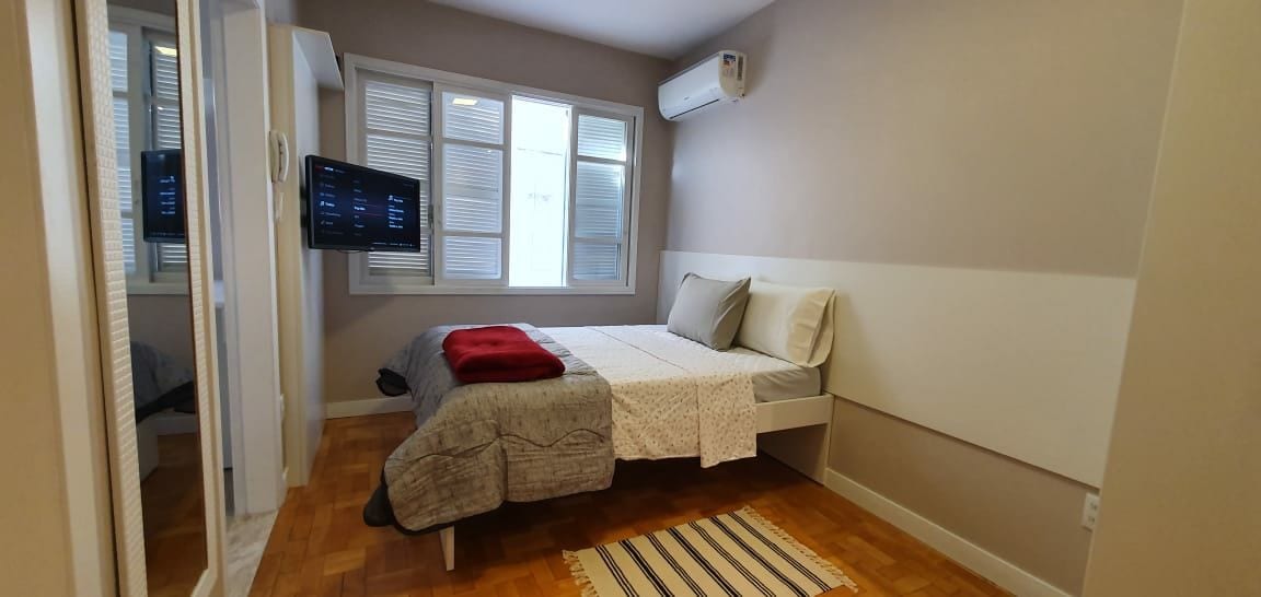 A modern bedroom features a spacious bed with light-colored bedding and decorative pillows. A soft rug lies on the wooden floor. Natural light enters through large windows, and a wall-mounted television is visible. An air conditioning unit is positioned above the bed for comfort.