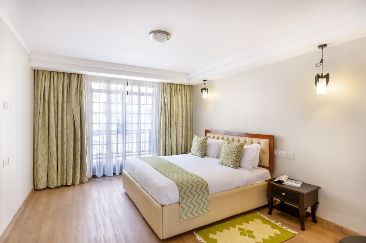 A spacious bedroom is presented with a large bed featuring decorative pillows. Soft curtains allow natural light to enter, framing a set of glass doors that lead to a balcony. A small table and decorative rug complete the serene atmosphere.