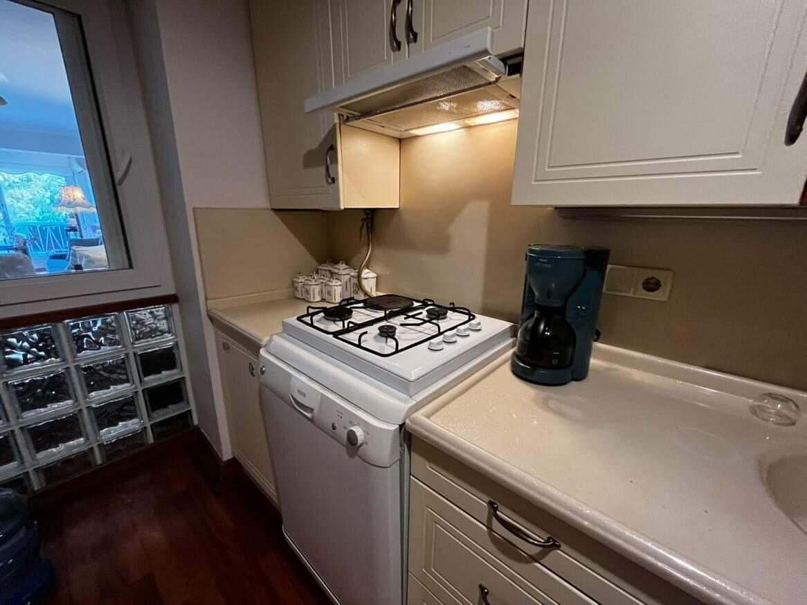 A well-organized kitchen is presented, equipped with a gas stove, white countertop, and a coffee maker. The cabinetry features light-colored finishes, and natural light filters in through a window, highlighting the clean design. A dishwasher and storage area are also visible.