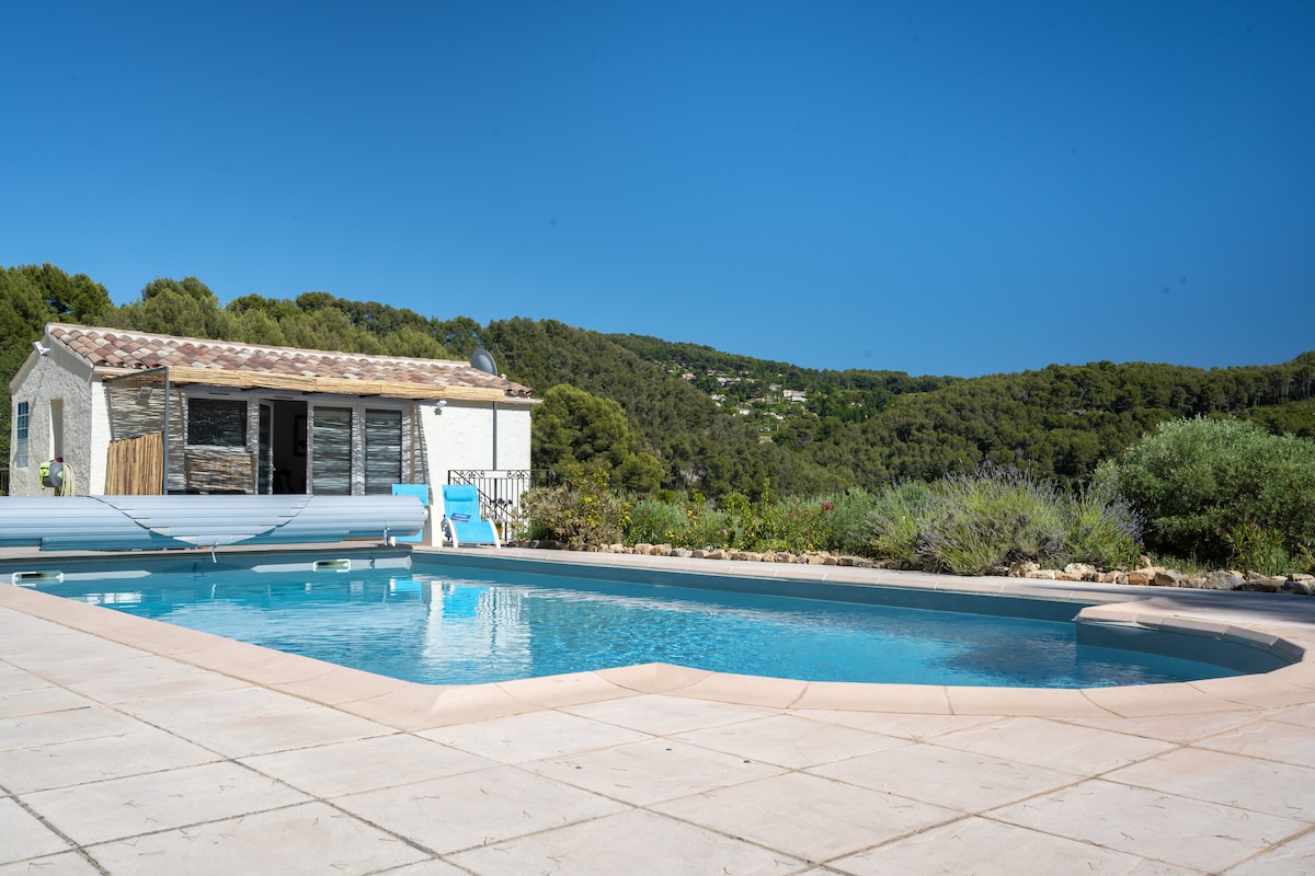 A serene pool area is visible, featuring a sparkling blue swimming pool surrounded by a tiled deck. A quaint building can be seen in the background, nestled among lush greenery. The landscape showcases gentle hills and trees, enhancing a peaceful rural setting.