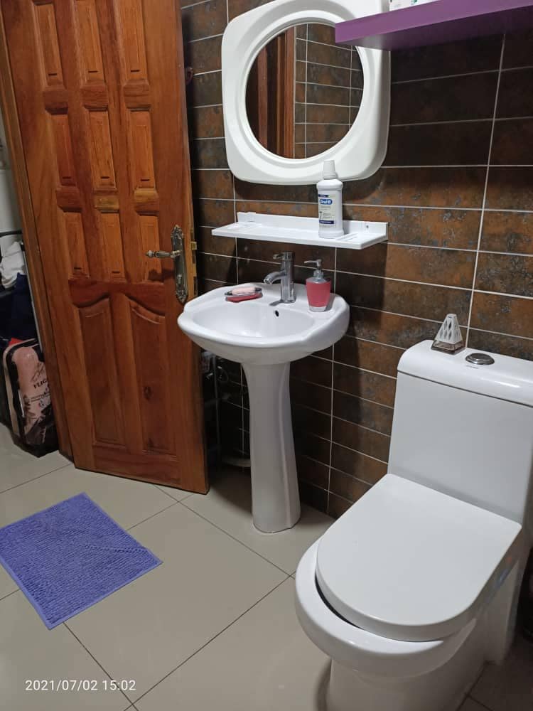 A clean bathroom features a wall-mounted mirror above a small shelf. A white sink is positioned beside a modern toilet. A bright purple mat is placed on the tiled floor, adding a touch of color to the space.