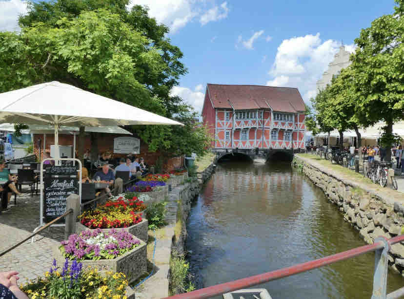 A scenic view of a historic building with timber framing stands by a calm canal. Colorful flower beds enhance the outdoor area, where people enjoy the waterfront. Shade is provided by large umbrellas, and bicycles line the pathway along the water's edge.