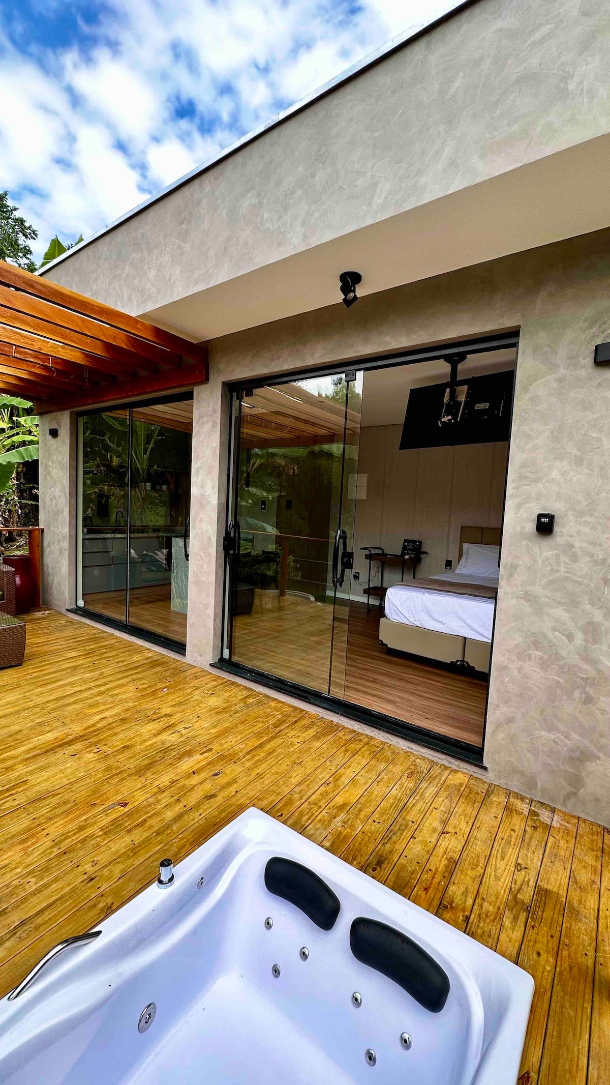 An outdoor deck with wooden flooring is showcased, leading to a glass door entrance. The modern cabin interior is visible, featuring a spacious bed and a dining area. Lush greenery surrounds the area, enhancing the serene ambiance.