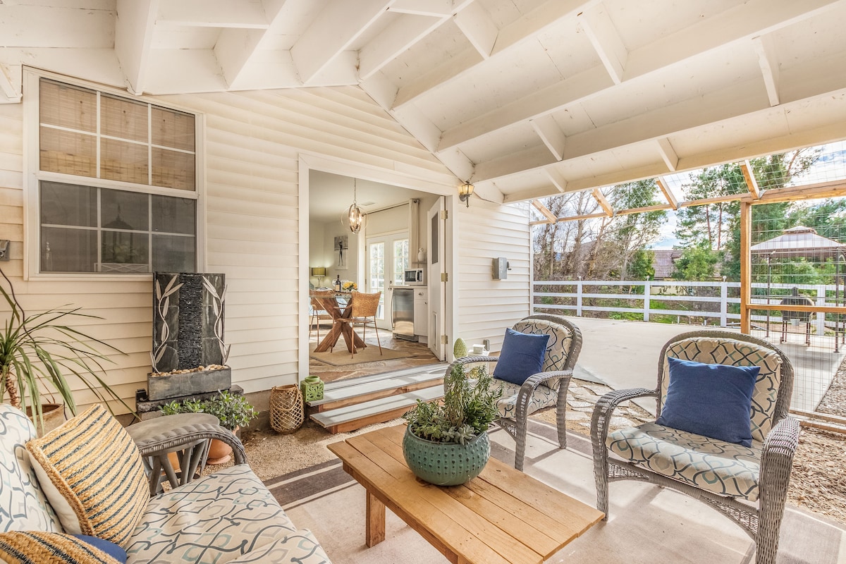 A cozy outdoor seating area features comfortable wicker chairs with decorative cushions, arranged around a wooden coffee table. Natural light filters through a covered porch, illuminating the space. A door in the background leads to an inviting dining area, with views of the surroundings outside.