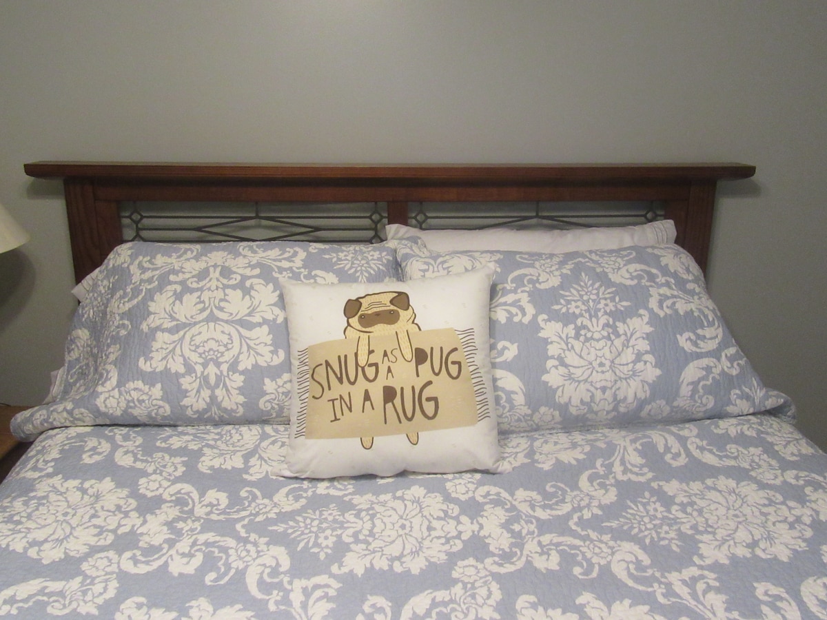 A neatly made bed is presented with soft blue-patterned bedding and white pillows. A decorative pillow featuring a playful illustration of a pug sits centered on the bed, adding a touch of character to the serene sleeping area.